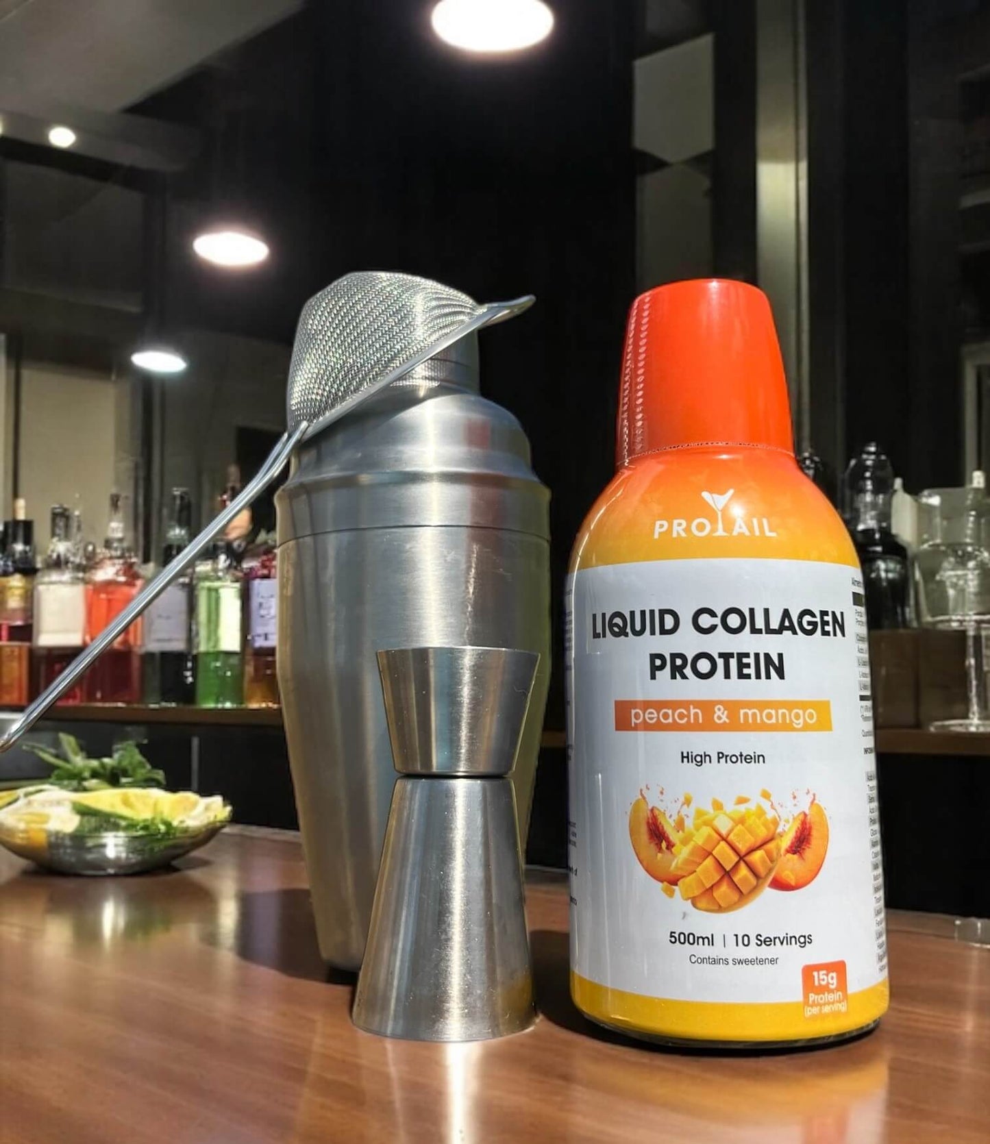Bottle of Protail Liquid Collagen Protein peach & mango flavor on a bar counter with a cocktail shaker.